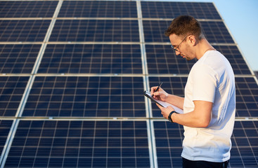 Side view of a young successful man standing by a solar pv plant looking in his folder pondering on...