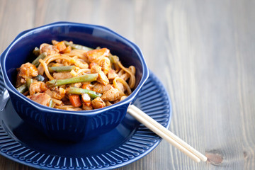 Teriyaki chicken with noodles and vegetables stir fry in a bowl, wooden background. Udon noodles, teriyaki chicken, vegetables, chopsticks. Closeup. Asian style dinner.