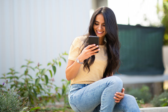 Smiling Young Woman Sitting Outside Looking At Cellphone