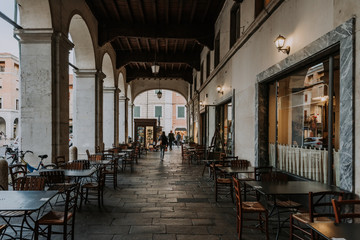 Fototapeta na wymiar Cozy narrow street with arches and tables of cafe in Ferrara, Emilia-Romagna, Italy. Ferrara is capital of the Province of Ferrara