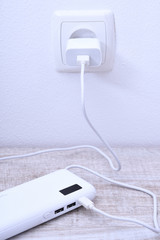 White phone power bank plugged in the socket on the wall for charging on wooden background