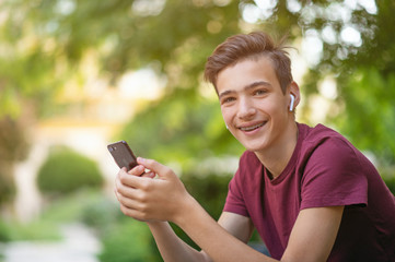 Naklejka na ściany i meble Happy teenage boy is using mobile phone, outdoors. Close-up portrait of a smiling young man with smartphone, in park. Cheerful teenager in casual clothes with cell phone in park. Soft focus