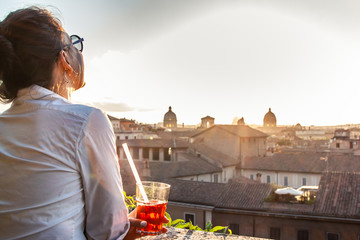 Young woman tourist fashion white dress with spritz cocktail in front of panoramic view of Rome cityscape from campidoglio terrace at sunset. Landmarks, domes of Rome, Italy.