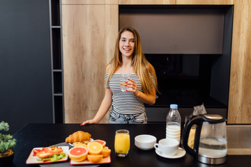 Beautiful young woman drinking water in kitchen with healthy food on table.