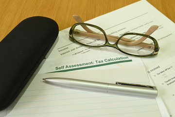 Close up view of a self assessment tax return form with a pen, a pair of glasses and a glasses case on a wooden table