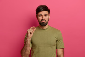 Dissatisfied bearded man makes little gesture, shows something tiny with fingers, purses lips,...