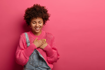 Lovely cheerful Afro American woman presses palms to heart, has grateful expression, thankful for received gift, closes eyes, wears fashionable clothes, isolated on pink wall, blank space aside