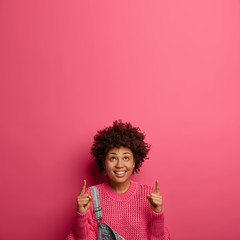 Cheerful optimistic female raises hands up, points above with positive grin, shows interesting promo, recommends good item, wears knitted jumper, isolated over rosy wall, copy space for your text