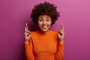 Happy dreamy woman sees something she wants very much, crosses fingers and hopes aspiring dream come true, makes wish and looks with great anticipation, wears casual orange jumper, stands indoor