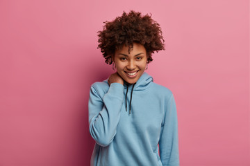 Shy romantic curly woman smiles happily, touches neck, looks directly at camera, enjoys spending free time in good company, listens postivie news, wears casual sweatshirt, has good mood, isolated