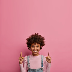 Wanna go there together and see. Positive dark skinned young woman points index finger above, advertises copy space for your information, smiles pleasantly, isolated over pink pastel background. - obrazy, fototapety, plakaty