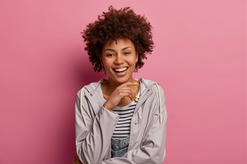 Obraz na płótnie Canvas Lovely optimistic woman with Afro hairstyle, wears casual anorak, touches chin gently, listens good news with pleasure, has carefree expression, isolated over rosy pastel wall, poses relaxed