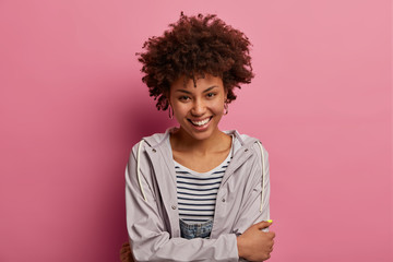 Happy cute woman with timid calm face expression, crosses hands over body, looks with gentle smile at camera, wears casual windbreaker, expresses positive emotions, isolated on pink background © WHstudio Leushin N