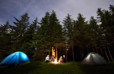 Tourist man showing his friends starry sky over the camping. The guys are sitting by fire and tourist tents on background of spruce forest and enjoying the evening sky. Outdoor recreation