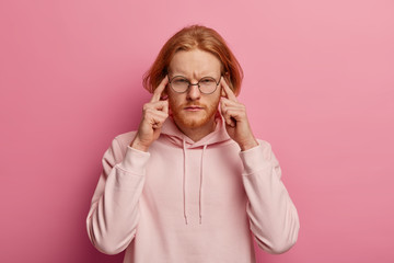 Portrait of intense redhead man presses index fingers on temples, focused on task, tries to concentrate on difficult task, has ginger thick beard, wears hoodie, isolated over pink background