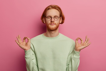 Portrait of relaxed ginger male tries to relax after hard work, meditates indoor, keeps hands in...