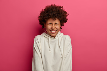 Portrait of sincere joyful teenage girl smiles broadly, squints face and has optimistic lifestyle, has funny expression, expresses good pleasant emotions, dressed in white sweatshirt, isolated on pink