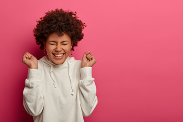 Successful upbeat woman with Afro hairstyle, clenches fists, has faith team win, celebrates success, smiles joyfully, wears white sweatshirt, says hooray, models over crimson wall, rejoices good luck