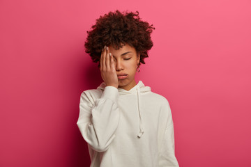 Fototapeta na wymiar Exhausted bored Afro woman covers half of face, sighs from tiredness, needs rest, has upset look, closes eyes, feels headache, dressed in white sweatshirt, poses over vibrant crimson background