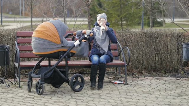 A young mother is sitting on a Park bench, rocking a stroller and drinking tea from a thermos. Family, parenthood concept - happy mother with stroller in park. Woman with baby outdoor.
