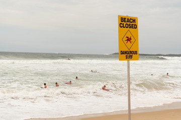 Beach closed sign at the beach. Despite the warning people are still swimming and surfing