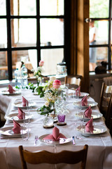A beautiful set table with pink napkins and roses in the front of a sunny window