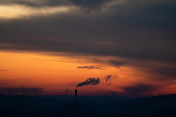 The silhouette of a smoking chimney in the far distance in front of a dramatic red sunrise and some wind turbines