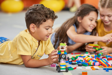 Fototapeta premium Kids playing block toys in playroom at nursery. Four children playing constructor while laying on the floor. Fun in kids room.