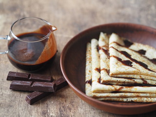 Pancakes poured with chocolate icing to the Shrovetide on a wooden brown background with a glass vase with chocolate icing. Place for text. Brown wooden background.