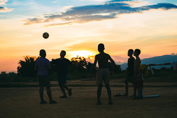 Fototapeta na wymiar Silhouette action sport outdoors of a group of kids having fun playing soccer football for exercise in community rural area under the twilight sunset. Poor and poverty children in development country.