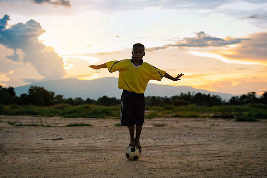 boy standing on a ball with bare foot while playing street soccer football for exercise in community rural area under the twilight sunset.