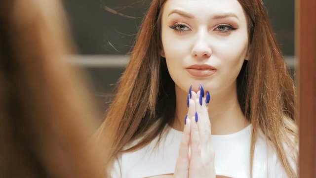 face of beautiful confident girl with long hair looking in the mirror in the interior, young brunette woman admiring herself makeup, female beauty concept