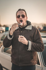 A man with a beard, sunglasses and a leather jacket is licking ice cream from a spoon. Cars seen parked in the background. Vintage parking ice-cream feast.