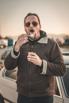 A Man With A Beard, Sunglasses And A Leather Jacket Is Licking Ice Cream From A Spoon. Cars Seen Parked In The Background. Vintage Parking Ice-cream Feast.