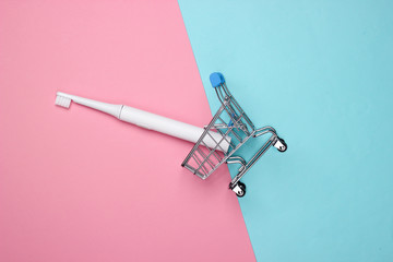 Mini shopping trolley with electric toothbrush on a pink-blue pastel background. Top view.