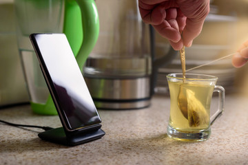 smartphone wireless charging on charging stand next to male hand make tea on kitchen tabletop