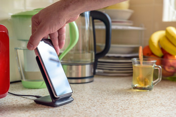 smartphone wireless charging on charging stand on kitchen tabletop. male hand placing phone to...