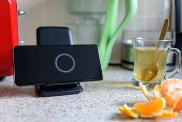 smartphone wireless charging on charging stand next to tea glass cup on kitchen tabletop