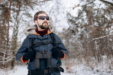 Enthusiastic hiker in anorak and mirrored glasses posing in snow-clad forest with binoculars on his neck © Kuz Production