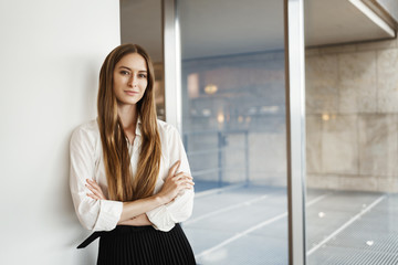 Successful young businesswoman lean wall and cross hands on chest, smiling confident and assertive, express readiness, just signed big financial deal for her company, standing inside office