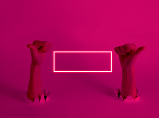 Female hands show thumbs up through the torn holes with pink light. Neon frame Creative art