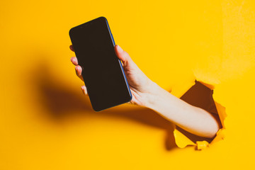 Female hand out of the hole in the paper wall, holding smartphone with a black screen.