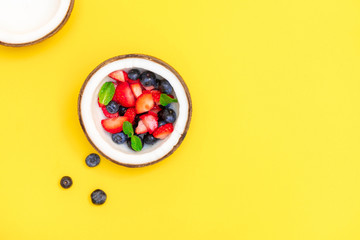 Directly above view of coconut bowl with fresh ripe berries on bright yellow background with copy space