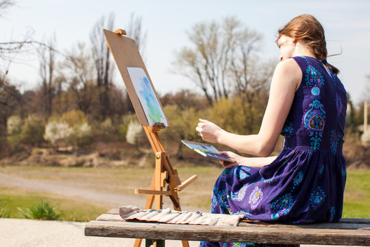 Young woman creative painter draws brush watercolor paints on canvas outdoors. Spring inspiration.