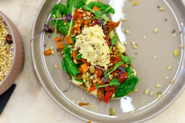 toast with homemade hummus and baked vegetables on non-yeast whole-grain bread, with spinach, baked peppers and carrots, a mix of seeds and micro greens on plate on the table