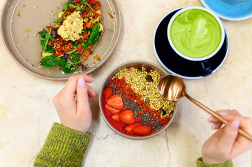 smoothie bowl with strawberry, goji, and green buckwheat granola, and matcha latte cup, and toast with hummus, and female hands with golden spoon, ready to eats. raw healthy food concept
