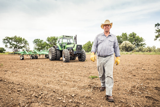 Farmer with cowboy hat walking across plowed field tractor in background. Bridger, Montana, USA