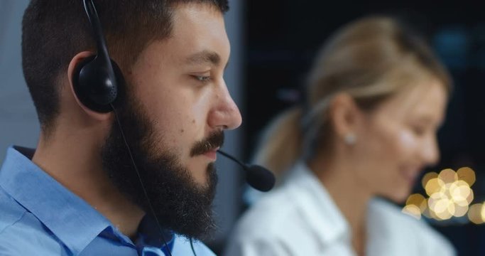 Close up of Caucasian handsome man in headphones and mic smiling and having videochat in call center. Male worker of call-center speaking with client in evening. Female co-worker behind.