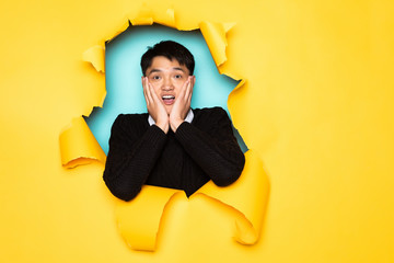 Young chinese man surprised keeps head in hole of torn yellow wall. Male head in ripped paper.