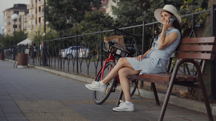 Fototapeta na wymiar A young woman sits on a bench in the city and talks on the phone, there is a bicycle next to her.